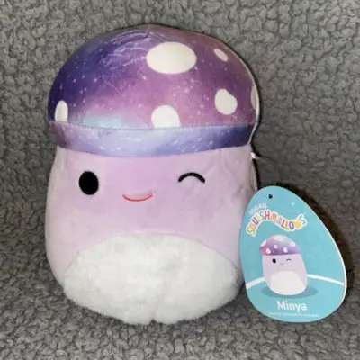 En lilla champignon squishmallow med en fuzzy hvid mave og en lilla kosmisk mønstret kasket med hvide pletter