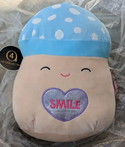 Une Squishmallow champignon avec un bonnet bleu tacheté de blanc et un cœur bleu disant sourire sur le ventre.