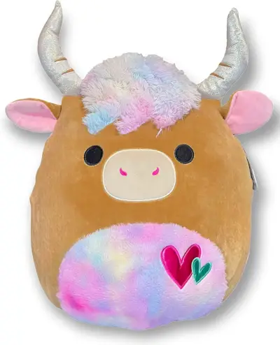 Squishmallow de vache brune avec des cornes brillantes, un ventre et une frange arc-en-ciel et des cœurs sur ce ventre