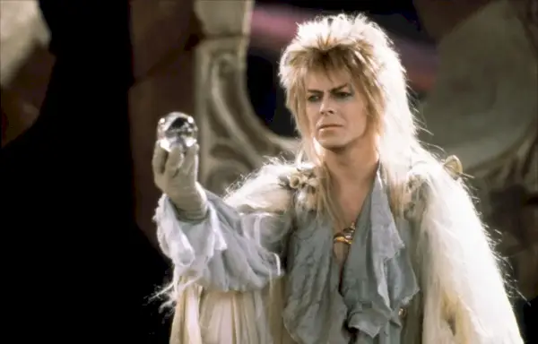 Jareth (David Bowie) pitää pystyssä lasipalloa, yllään kiiltävä hopeaasu ja vahva meikki.