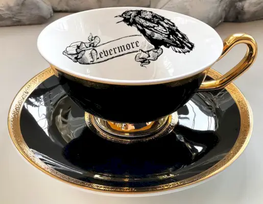 Une tasse à thé noire et une soucoupe avec une poignée et des bords dorés, ainsi qu'un dessin au trait d'un corbeau et le mot Nevermore imprimé à l'intérieur.