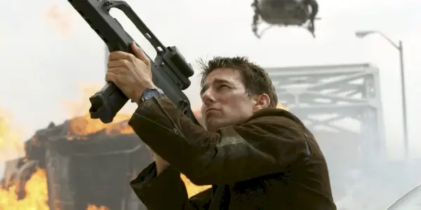 Tom Cruise som Ethan Hunt i MIssion: Impossible III, peker en pistol opp mot himmelen med et helikopter og eksploderende kjøretøy i bakgrunnen.