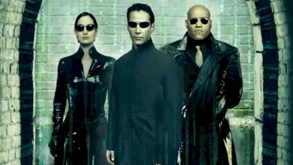 Neo (Keanu Reeves), Trinity (Carrie-Anne Moss) in Morpheus (Laurence Fisburne) stojijo skupaj in strmijo v kamero v temnih sončnih očalih na plakatu The Matrix Reloaded.