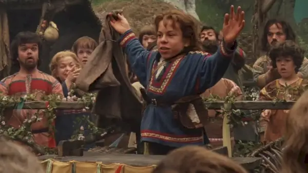 Warwick Davis como Willow en la película Willow levantando los brazos triunfalmente entre sus compañeros del pueblo.