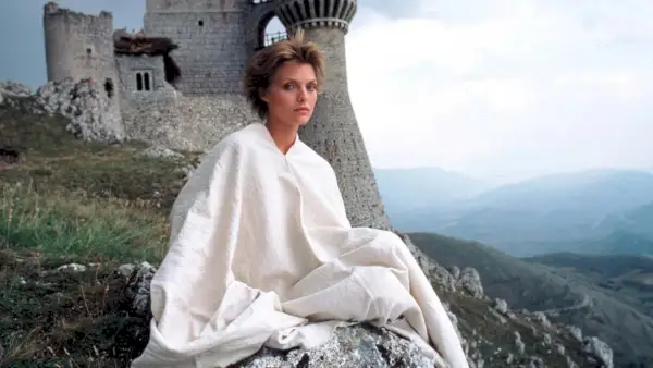 Michelle Pfeiffer como Isabeau en la película Ladyhawke, sentada en una ladera rocosa en el centro de Italia, con un castillo medieval al fondo.