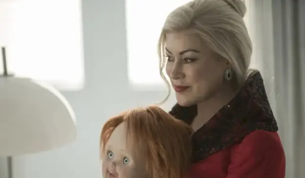 Eine blonde Frau (Jennifer Tilly) trägt einen roten Mantel und hält die Chucky-Puppe auf ihrem Schoß