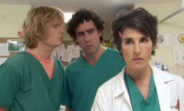 Fotograma de Green Wing; Julian Rhind Tutt, un hombre blanco de cabello rubio, está junto a Stephen Mangan, un hombre blanco de cabello oscuro. Ambos vistiendo batas verdes. Frente a ellos se encuentra Tamsin Greig, una mujer blanca de cabello oscuro, con una bata blanca de médico.