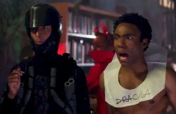 Fotograma del episodio Epidemiología de Community; Danny Pudi, vestido todo de negro con casco de motociclista y tubos en el torso, junto a Donald Glover, sin camisa y con un cuello de papel higiénico con la inscripción Drácula. Donald Glover está gritando.