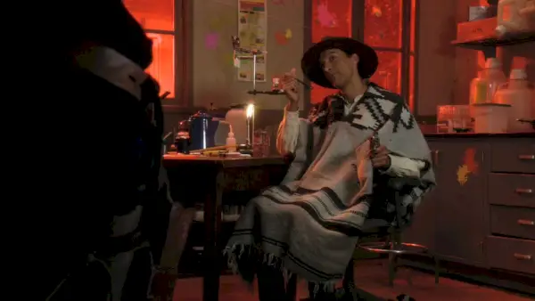 Siéntese del episodio de la comunidad Un puño lleno de bolas de pintura; Danny Pudi, con un sombrero de vaquero negro y un poncho gris, está sentado en una mesa de laboratorio comiendo frijoles en lata.